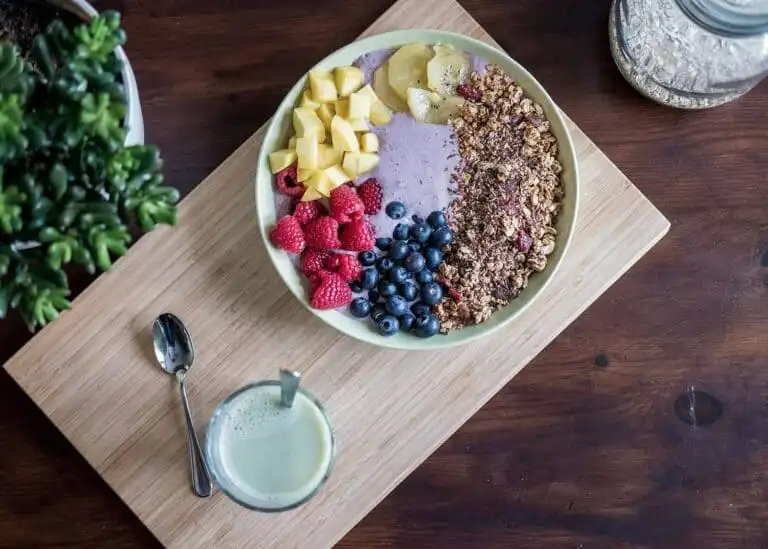 Fresh fruit smoothie bowl with berries, mango, pineapple, granola, and a side of green juice on wooden table.